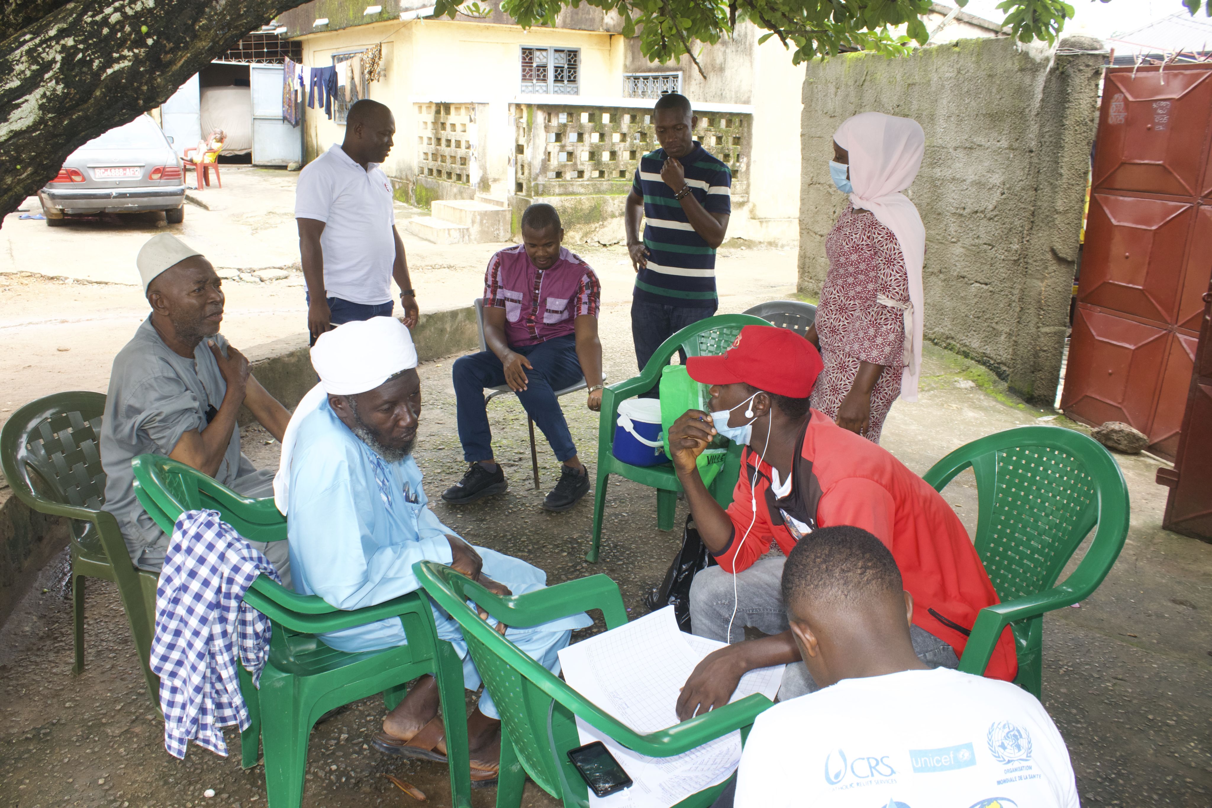 men sitting outside on plastic chairs and talking, and men and woman standing and observing