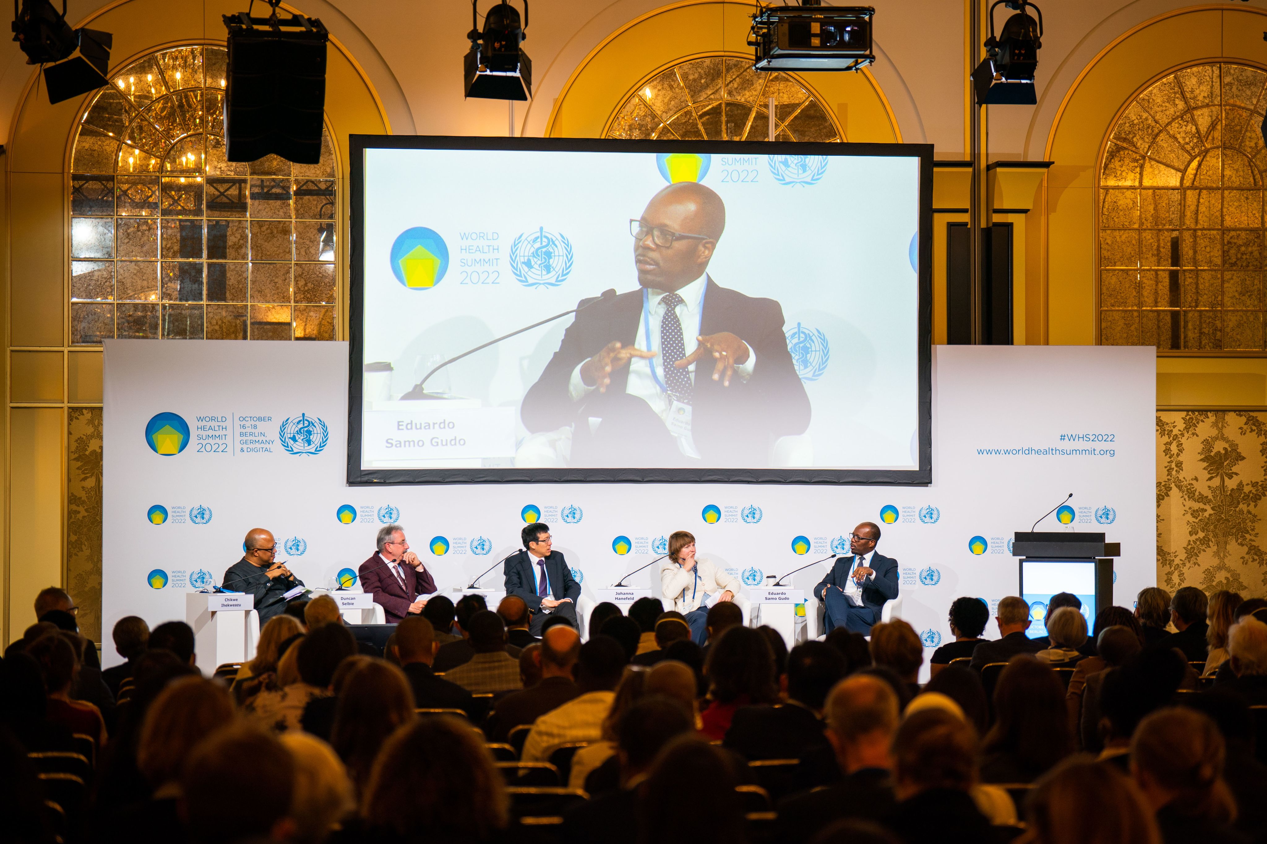 four men and one woman on a stage with audience in the room, one man is talking